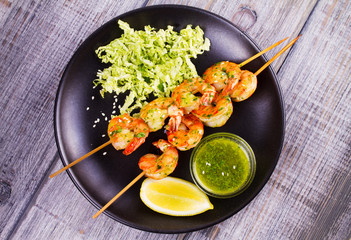 Cilantro grilled shrimps on skewers with sesame napa cabbage, green butter sauce and lemon. Skewered prawns on black plate. View from above, top studio shot