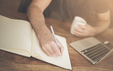 man at rustic wooden desk taking notes on pad while working on a laptop computer