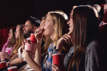 Big group of young best friends, stylish girls and boys watching funny film at cinema, spending free time together, eating tasty popcorn. Pretty and stylish women drinking cola, looking at big screen. - obrazy, fototapety, plakaty