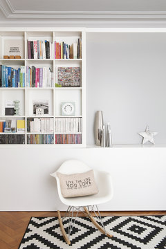 Salon et biblioth&egrave;que avec chaise &agrave; bascule design blanche, tapis &agrave; motif noir&blanc, et vases argent&eacute; et livres d'art
