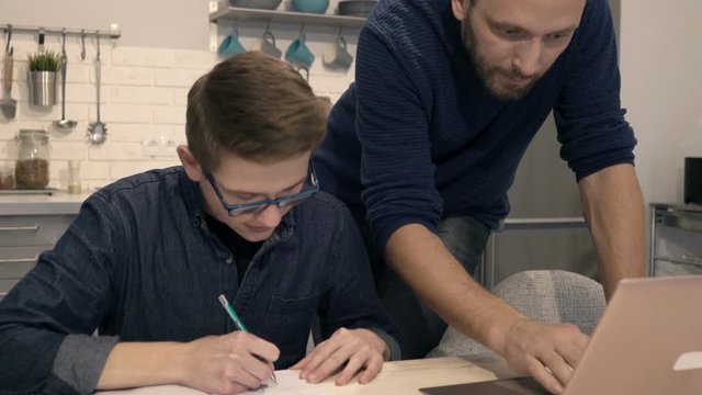 Father with laptop helping his son doing homework in kitchen at home
