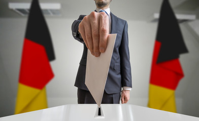 Election or referendum in Germany. Voter holds envelope in hand above ballot. German flags in...
