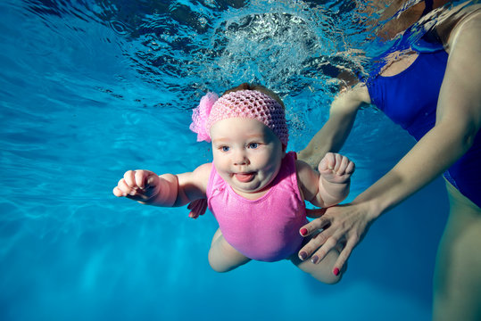 Little an infant girl swims underwater in the pool with open eyes, in a pink dress and mom helps her. Portrait. Close-up. Bottom view from under the water. Landscape orientation