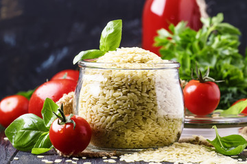 Italian uncooked pasta riso or orzo in a glass jar, tomatoes and green basil, black background,...