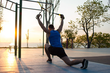 Fototapeta na wymiar workout with suspension straps In the outdoor gym, strong man training early in morning on the park, sunrise or sunset in the sea background 
