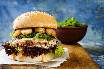 Asian style salmon burger with grilled shrimps, seaweed, lettuce and spicy sriracha mayo sauce served on pieces of brown paper on a rustic wooden board. Bright blue background with copyspace