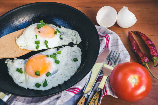 Fried eggs on pan, tomato and chili pepper on wooden background