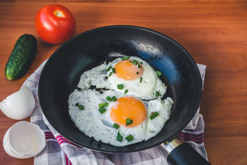 Fried eggs in pan, cucumber and tomato on wooden background