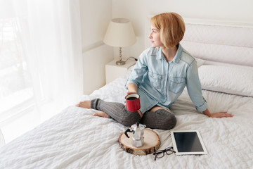 Young woman looking at window while drinking coffee sitting on bed in bedroom in the morning