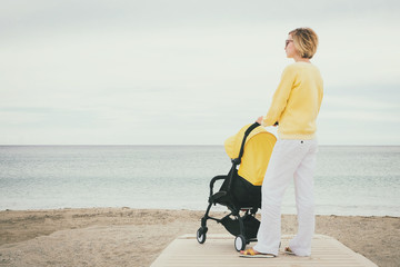 Young mother looking through the sea while walking with baby stroller at the beach