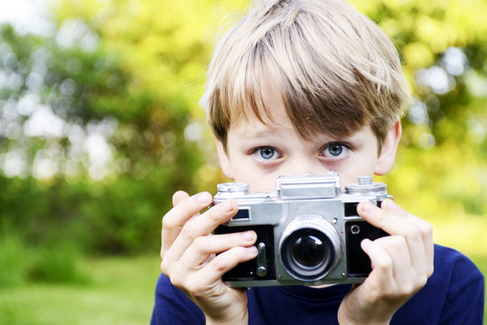 Little child blond boy with an old camera shooting outdoor. Kid taking a photo using a vintage retro film camera. Summer nature background