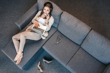 Overhead view of woman resting on couch and talking on smartphone with laptop on her knees.