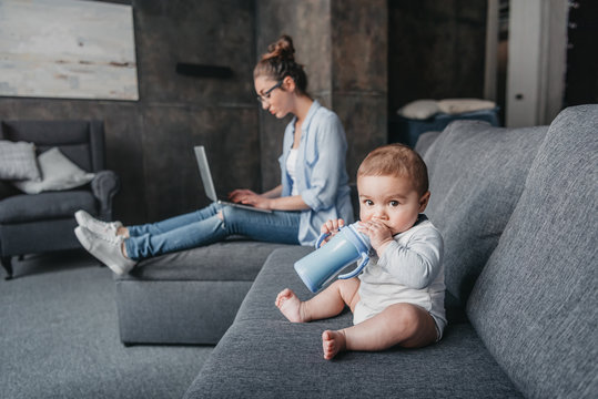 Adorable little boy drinking milk from baby bottle on couch while his mother working on laptop at home. family life concept