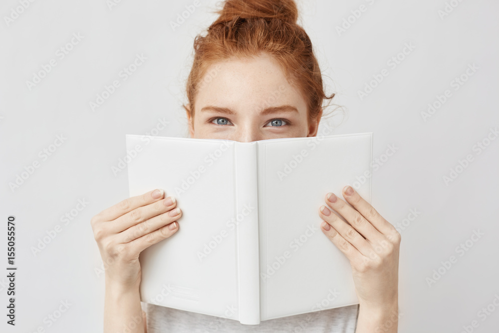 Wall mural Portrait of ginger girl hiding behind book looking at camera.