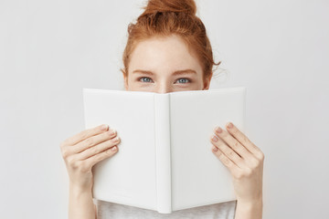 Portrait of ginger girl hiding behind book looking at camera.
