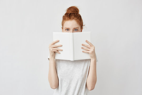 Redhead girl hiding face behind notebook looking at camera frightenedly.