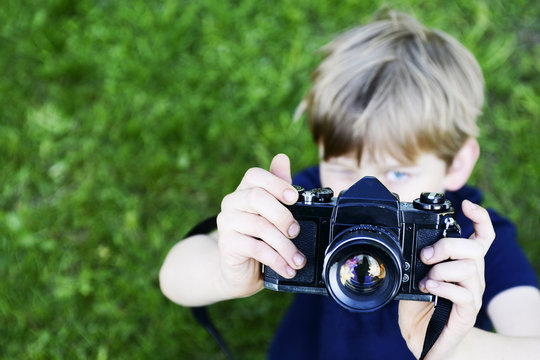 Little child blond boy with an old camera shooting outdoor. Kid taking a photo using a vintage retro film camera. Green summer grass lawn background.