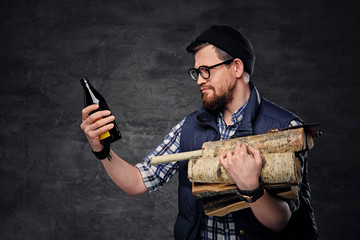 A drunk bearded male dressed in fleece shirt holds firewoods in his arms and drinks craft bottled...