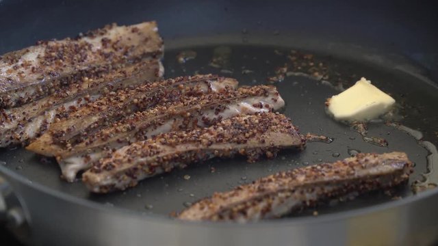The man throws butter on the pan while cooking meat. The meat is spiced with pepper and herbs.