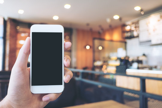 Mockup image of hand holding white mobile phone with blank black screen in modern loft cafe