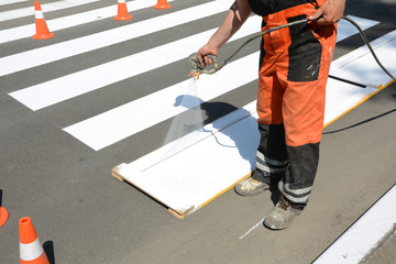 Worker is painting a pedestrian crosswalk. Technical road man worker painting and remarking pedestrian crossing lines on asphalt surface using paint sprayer gun.