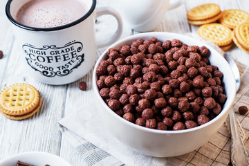 Hearty and tasty breakfast for children, cornflakes, chocolate balls with milk, cocoa and biscuits on a light wooden background 