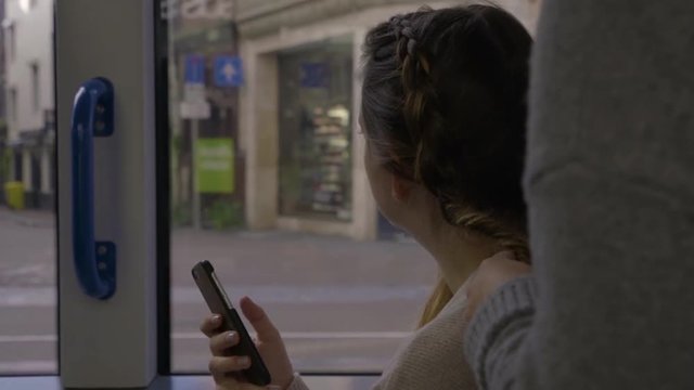 Young Woman Holds Smartphone And Looks Out The Window, While Her Friend Braids Her Hair, On The Light Rail In Amsterdam, Netherlands