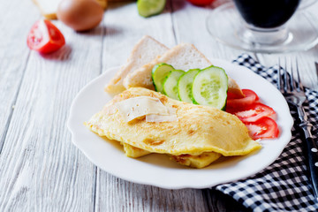 Hearty and tasty breakfast, traditional in the hotel, omelette from chicken eggs with cheese, fresh vegetables - cucumber and tomato, white toast and black coffee on a light wooden background