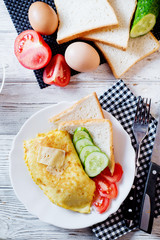 Hearty and tasty breakfast, traditional in the hotel, omelette from chicken eggs with cheese, fresh vegetables - cucumber and tomato, white toast and black coffee on a light wooden background