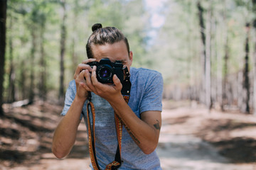 Young hipster millenial with man bun makes photograph on vintage analog film camera in middle of adventure trip through wild forest