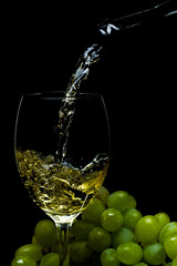 White wine pours from a bottle into a glass, a bunch of grapes, a black background, studio light