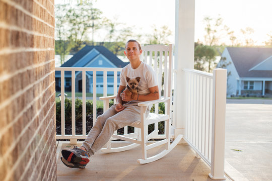 Cool and casual young man sits on the porch on rocking chair with his best friend puppy pet Yorkshire Terrier on his lap in the american house on sunset