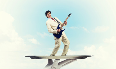 Businessman on metal tray playing electric guitar against blue sky background
