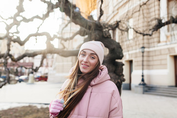 The beautiful girl walks during a winter time. On her a gentle-pink cap and a jacket. The girl smiles and looks at us. Easy beams of the sun fall on her hair and a face.