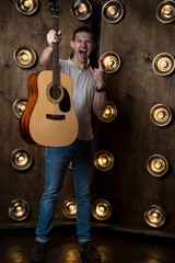 Guitarist, music. A young guy is standing with an acoustic guitar in his hand, in the background with lights behind him. Vertical frame