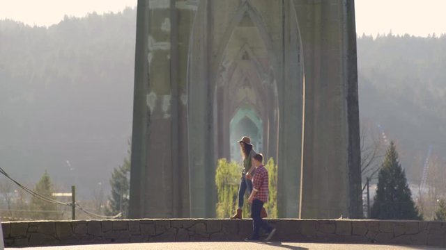 Couple Walk Under Beautiful St Johns Bridge, Young Woman Jumps Into Her Boyfriends Arms And He Twirls Her Around In His Arms