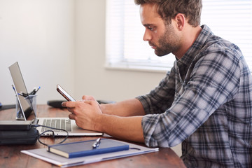 Young adult caucasian man busy using his cellphone while seated at his beautiful wooden desk at home, wearing a checked shirt.