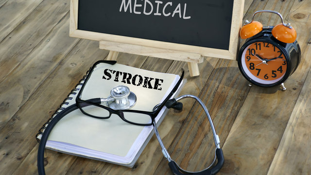 notebook with the words "STROKE" and stethoscope, glasses, chalk board, alarm clock on a wooden table. medical and healthcare concept.