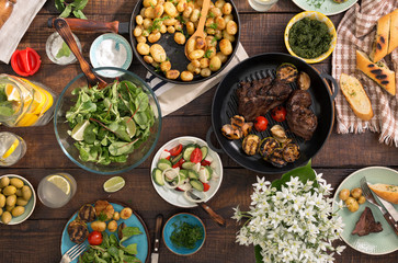 Dinner table with grilled steak, vegetables, potatoes, salad, snacks, lemonade