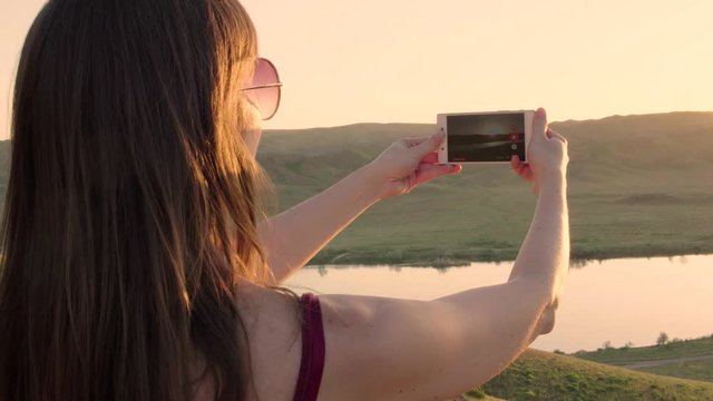 Close up from the back of girl or woman taking photo of sunset lake view landscape with her iphone