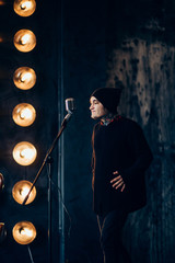 beautiful male hipsters in tattoos in black hat, singing a song, holding the microphone, emotionally, with feeling, on a dark background with bright lights on the wall