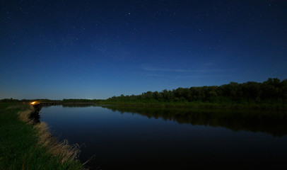 The stars in the night sky are reflected in the river. The landscape is photographed by moonlight.