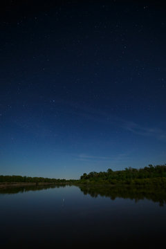 The stars in the night sky are reflected in the river. The landscape is photographed by moonlight.