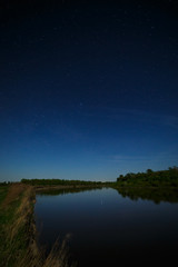 The stars in the night sky are reflected in the river. The landscape is photographed by moonlight.