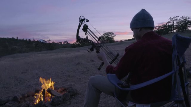 Man Sits In Camp Chair By Fire, At Sunset, And Checks Out His Hunting Bow