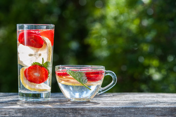 A glass and a bowl of detox water. Strawberry, lemon and mint with cool clean water,  backdrop of a green garden. Detox, diet, Cookery, sports,
Proper nutrition, sports nutrition, body cleansing.

