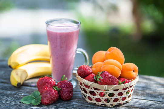 glass with strawberry apricot smoothies on a wooden table, a background of a green garden. A basket with strawberries and apricots, bananas near