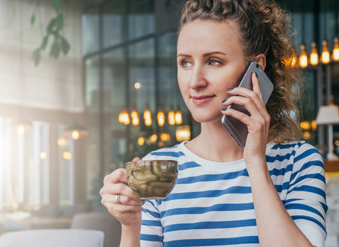 Young cheerful businesswoman in striped T-shirt is sitting at table in cafe, drinking coffee and talking on cell phone.