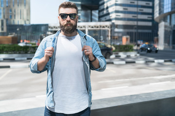 Bearded hipster man in sunglasses, dressed in white T-shirt, stands on city street.Mock up. Space for logo, text, image.