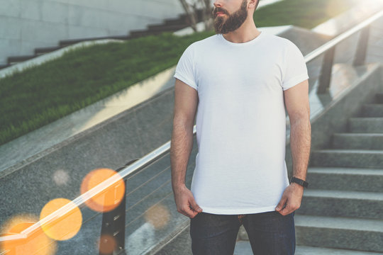 Summer day. Young bearded hipster man dressed in white t-shirt is stands on steps. On background is lawn. Mock up.